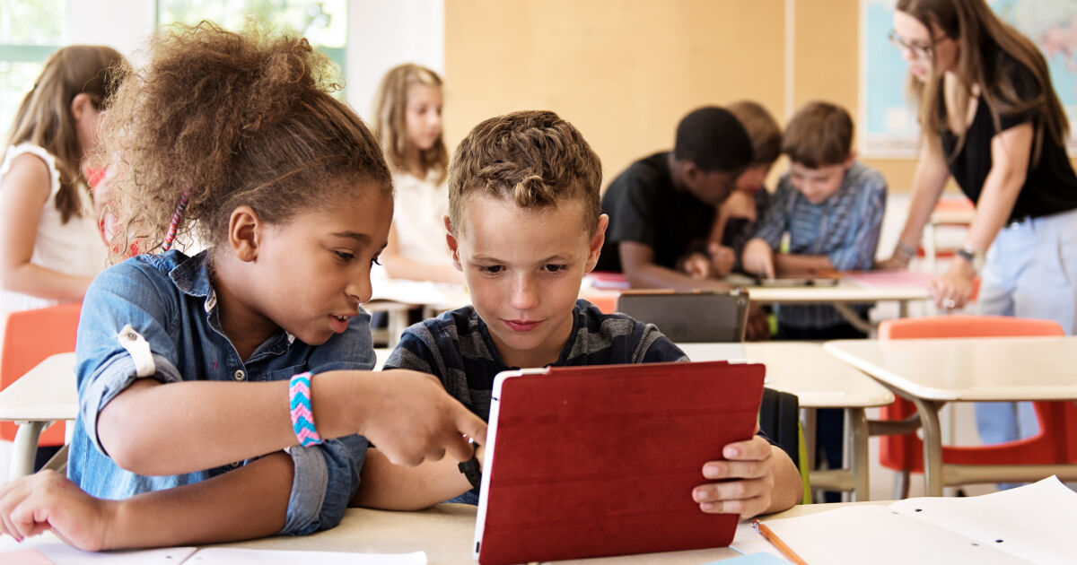Two young students - about middle-school age - sit next to one another at a desk. The child on the right - a boy - reads an iPad. The child on the left - a girl - points at the iPad screen and reads along with the boy.